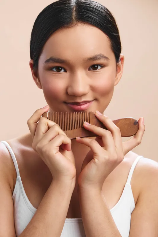 Asian woman with dark hair gently bites a wooden comb in a soft studio setting, creating a candid moment of selfcare and natural beauty with a calm, inviting vibe.