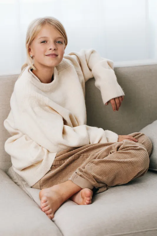 Young girl sits on a light sofa wearing a soft cream sweater and corduroy pants, relaxed and comfortable, creating a warm lifestyle portrait with natural, inviting lighting.