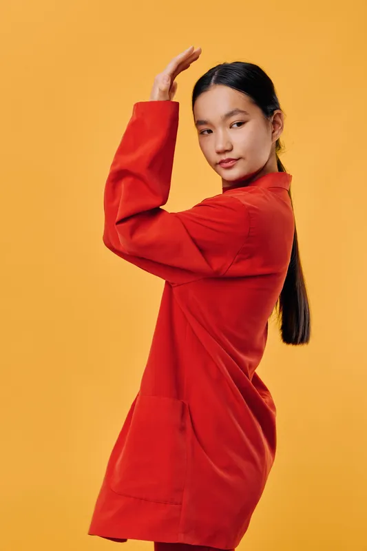 Young woman standing sideways with long dark hair wearing a red jacket posing with raised arm against yellow background in studio. Fashion, casual, confident, stylish look.