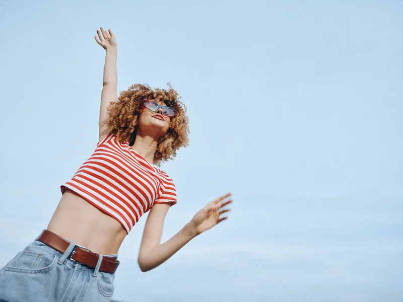 A joyful young woman with curly hair in a red striped crop top and jeans spins against a vast blue sky, capturing carefree summer energy and movement.
