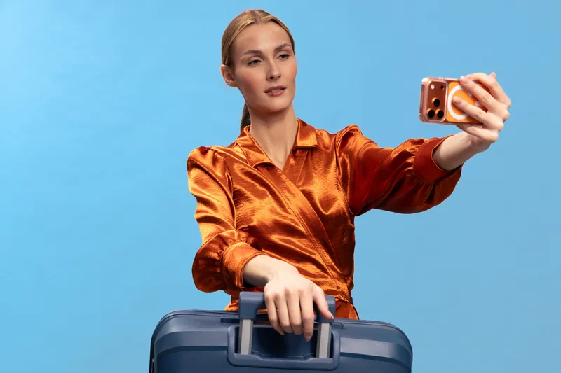 Young woman in orange satin blouse taking selfie with smartphone while holding blue suitcase on blue background. Concept of travel, vacation, tourism, and modern lifestyle.