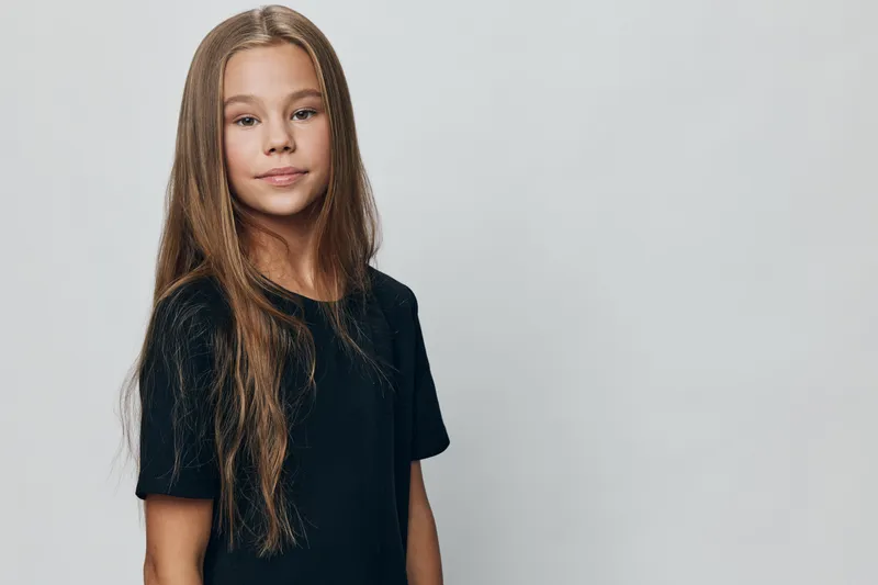 Girl with long hair in black t shirt standing against light grey background looking at camera with calm confident expression and natural pose in studio.