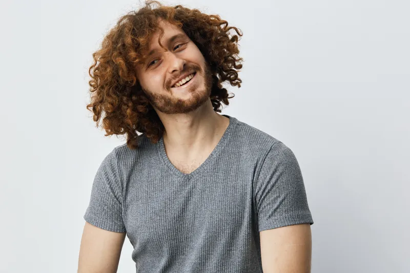 Happy young man with curly hair smiling in casual gray shirt against a light background, showcasing confidence and positivity