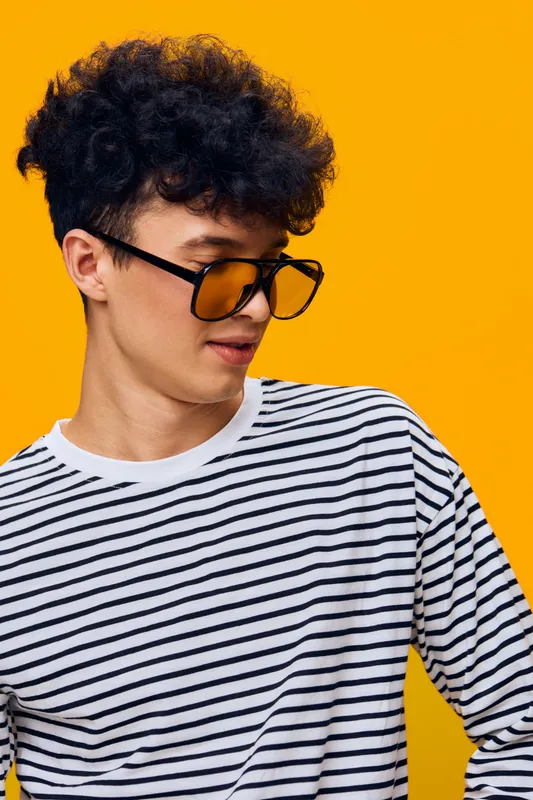 young man with curly hair wearing striped shirt and stylish orange tinted sunglasses on bright yellow background, casual fashion portrait, vibrant colors