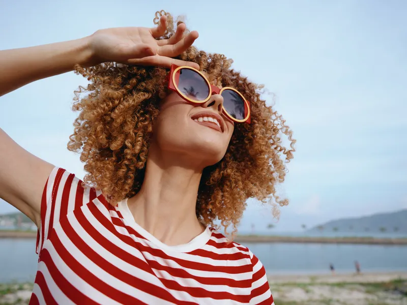 A smiling woman with curly hair wearing round sunglasses and a red striped shirt stands on a sunny beach, radiating carefree summer vibes and style.
