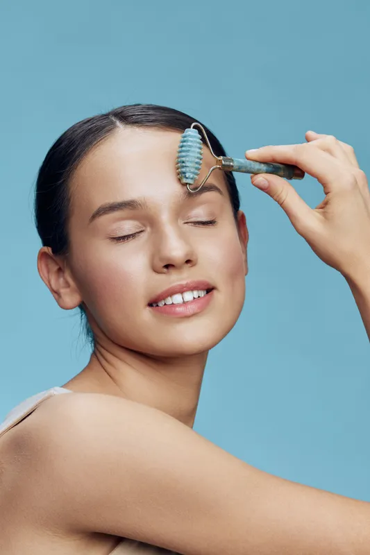 Young woman enjoying a facial roller treatment, smiling with eyes closed against a light blue background, promoting skincare and relaxation, beauty concept