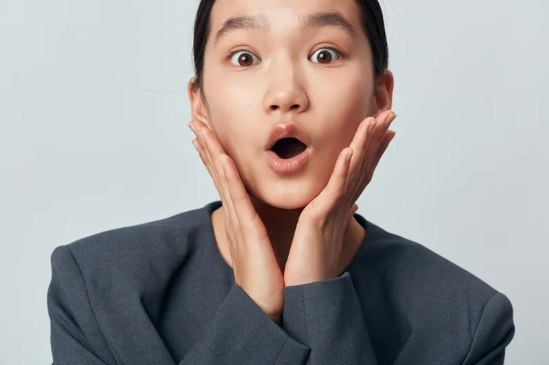 Woman with shocked expression and open mouth holding face with hands for surprise emotion on gray background. Female in gray jacket showing disbelief or amazement closeup.