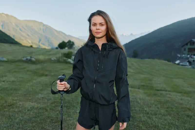 A confident young woman stands outdoors in a scenic mountain meadow, holding a trekking pole. She wears a dark jacket and shorts, looking ahead with calm determination, embodying adventure and
