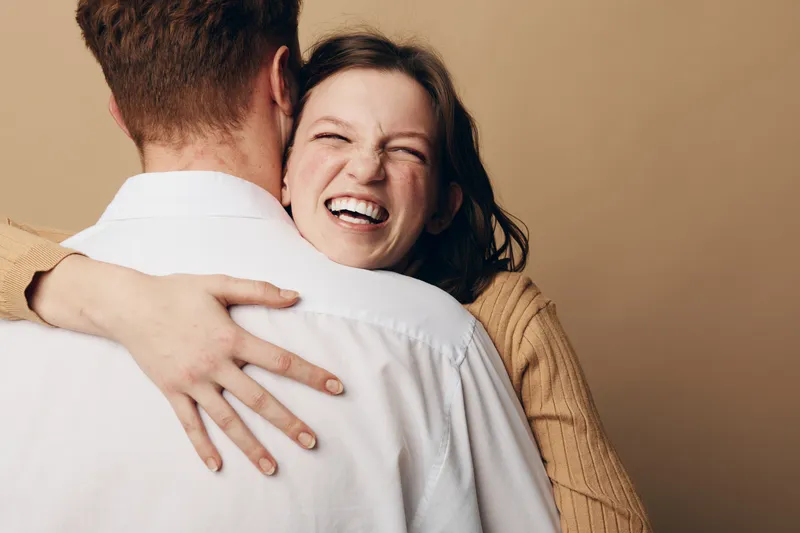 Happy woman with brown hair hugs man wearing white shirt, smiling with closed eyes in warm beige studio. Concept of love, affection, relationship, and joyful moment between couple.