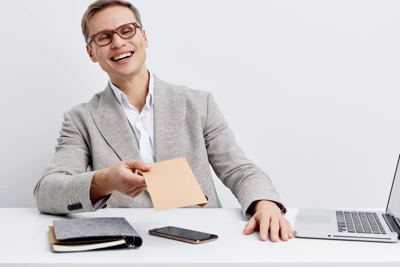 Bright smiling young Caucasian man in gray blazer and white shirt sitting at desk with laptop, notepad, and smartphone smiling and holding a small envelope isolated on white background studio portrait
