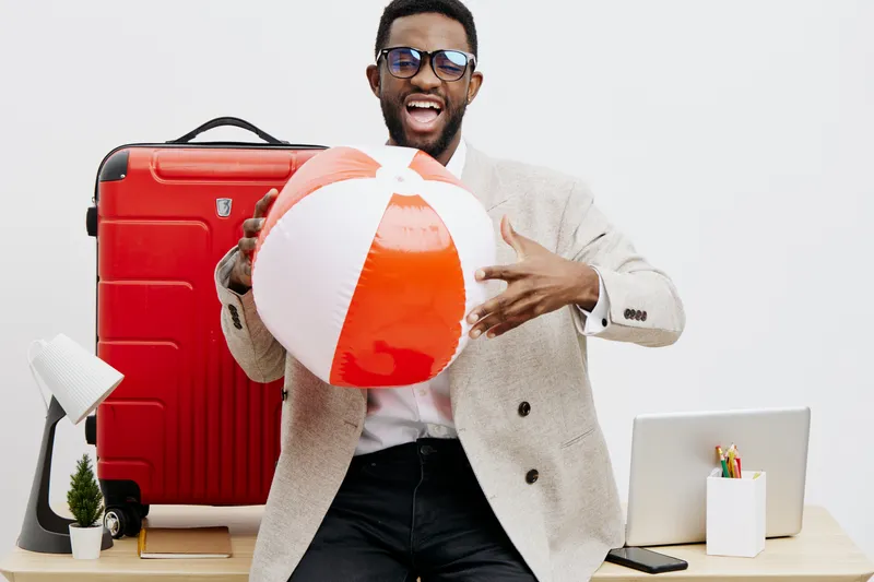 Joyful young African American man in casual clothing holding a beach ball, standing in front of travel essentials on desk, including a red suitcase, laptop, and small plant, bright indoor setting, lif