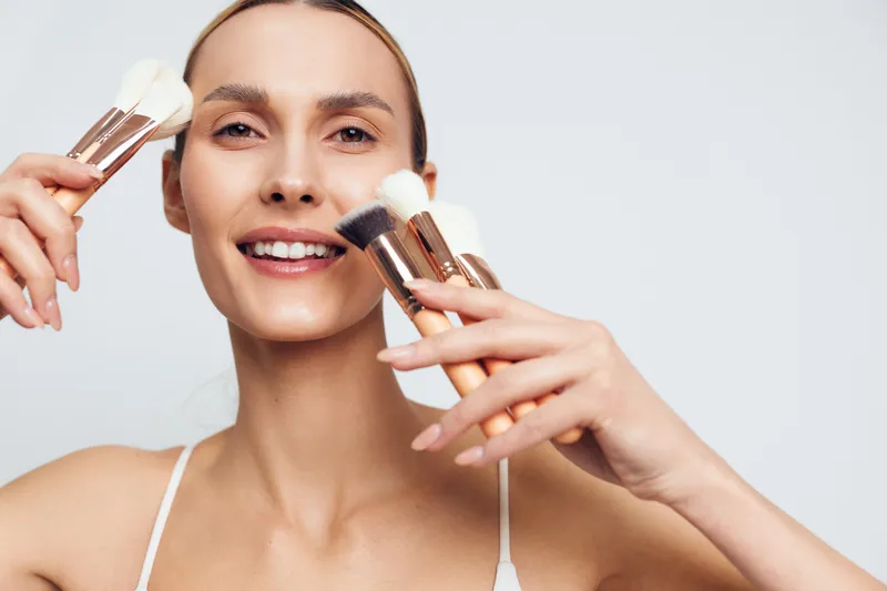 A smiling young woman applies makeup with brushes in a bright studio, showcasing a playful beauty routine and confident skincare moment.