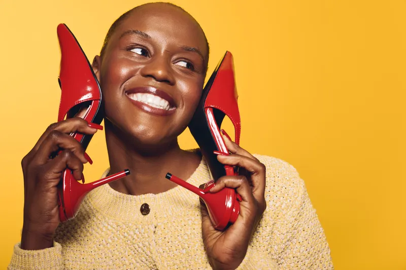 Smiling black woman with dark skin holds red high heels near face against yellow background. Concept of fashion, beauty, joy, and confident lifestyle with vibrant colors and happy mood.