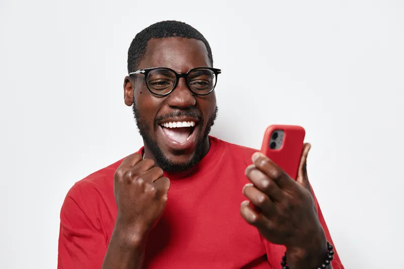 African American man with dark skin wearing glasses and red shirt smiling excitedly while looking at smartphone, celebrating success or good news indoors on white background.