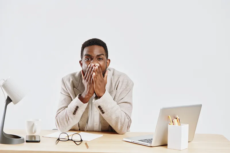 Portrait of surprised young man with dark skin, sitting at desk in bright modern office with laptop, eyeglasses, and stationery on table. Male expression of shock or disbelief. Studio shot with neutra