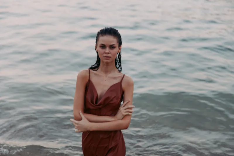 Portrait of a woman standing in shallow coastal water, wearing a burgundy dress. She gazes softly at the camera with a calm, confident expression as gentle waves lap at her waist.
