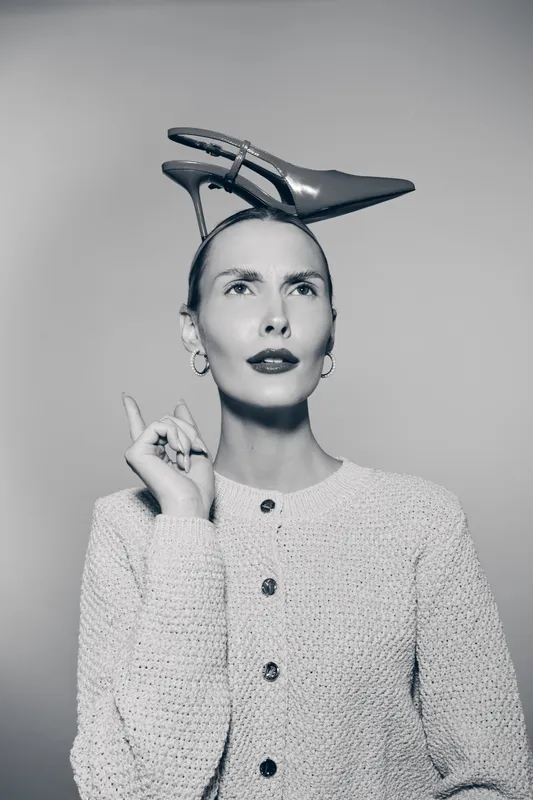 Caucasian white woman with fair skin wears high heel shoe on head and looks thoughtful, dressed in knitted sweater. Concept of fashion and creativity.