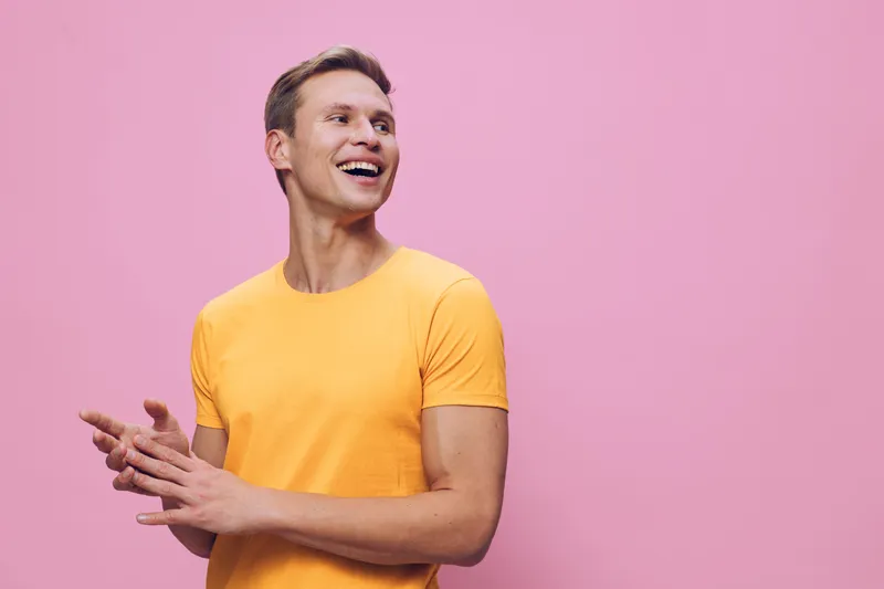 Cheerful smiling young man with light skin wearing a bright yellow t-shirt, standing against a pink background. His expression is joyful and relaxed, capturing a positive and vibrant mood in a casual 