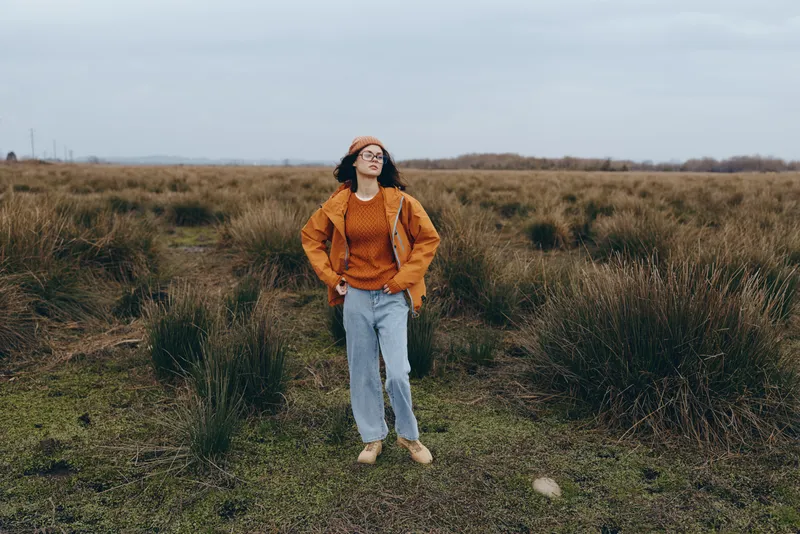 Woman lifestyle outdoors in a grassy field, smiling while walking along a path. She wears a brown jacket, orange sweater, jeans, beanie and sunglasses. Overcast sky, soft natural light.