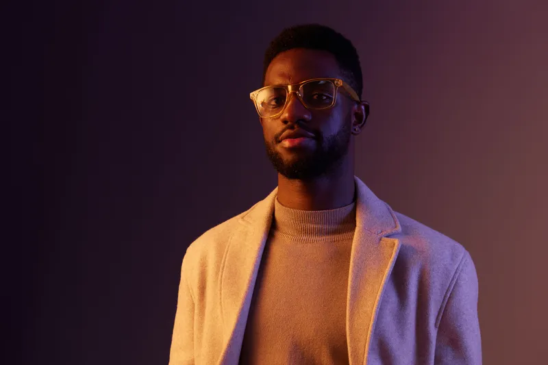 Stylish young man in glasses with a neutral expression, wearing a turtleneck sweater and a light overcoat, set against a colorful gradient background