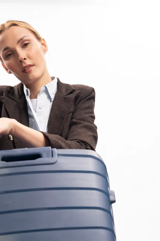 Young woman in a brown jacket leans on a large blue suitcase with a serious expression. Travel concept with luggage, business style, and preparation for a trip on white background.