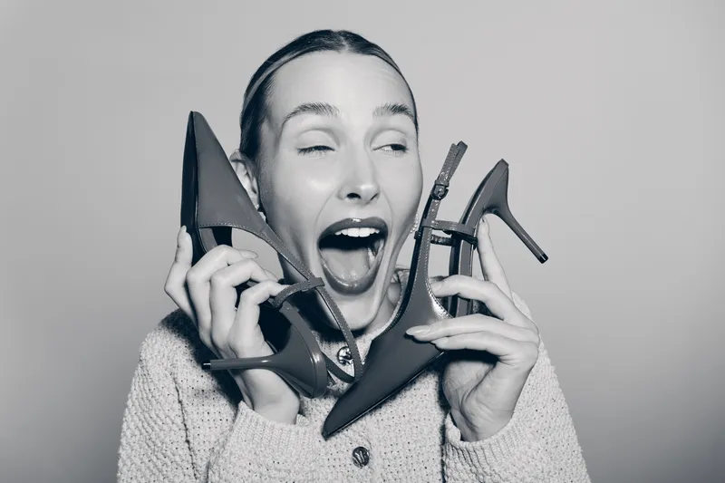 Young white woman with light skin holding two high heel shoes near her face, expressing excitement and joy in studio.