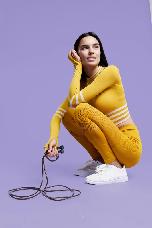 Woman sport fitness dressed in bright yellow athletic wear squats holding a jump rope, demonstrating health, strength, and focus against a solid lavender backdrop.