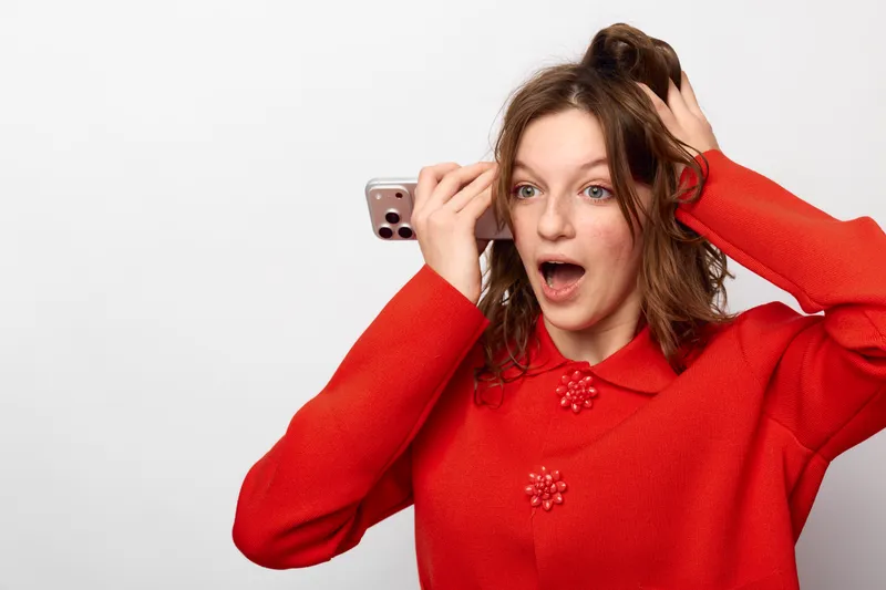 Surprised young woman in red sweater holds smartphone near ear with open mouth and wide eyes on white background. Concept of unexpected news, shock, and communication.