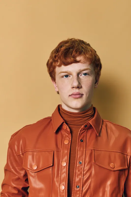 Young man with red hair wearing brown leather jacket and turtleneck posing against beige background. Studio portrait with casual confident expression and stylish outfit for fashion concept.