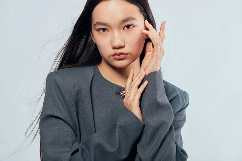 Young woman with long black hair wearing gray sweater posing with hand near face on gray background for fashion portrait or beauty and style concept in studio.