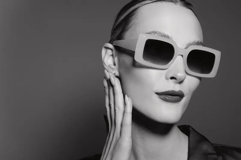 Stylish Caucasian woman with fair skin wears oversized sunglasses and red lipstick posing with hand near face on grey background. Fashion portrait with modern accessories and elegant look.