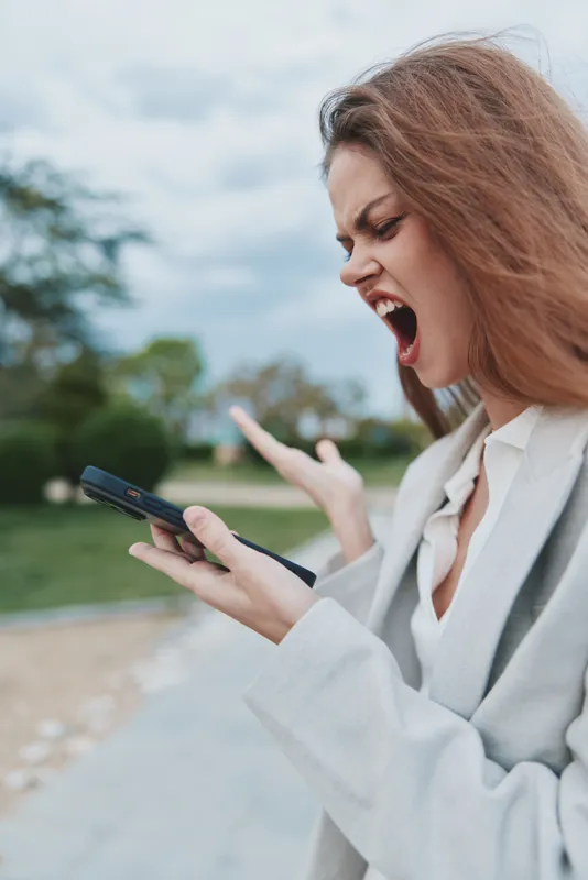 Angry woman shouting in frustration at mobile phone outdoors, Caucasian female expressing stress and emotional tension with blurred green park background