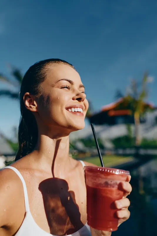 Vibrant outdoor portrait of a woman enjoying a refreshing smoothie under bright sun, relaxed posture, tropical backdrop, clear blue sky, summer mood, natural colors, confident smile, juice glass