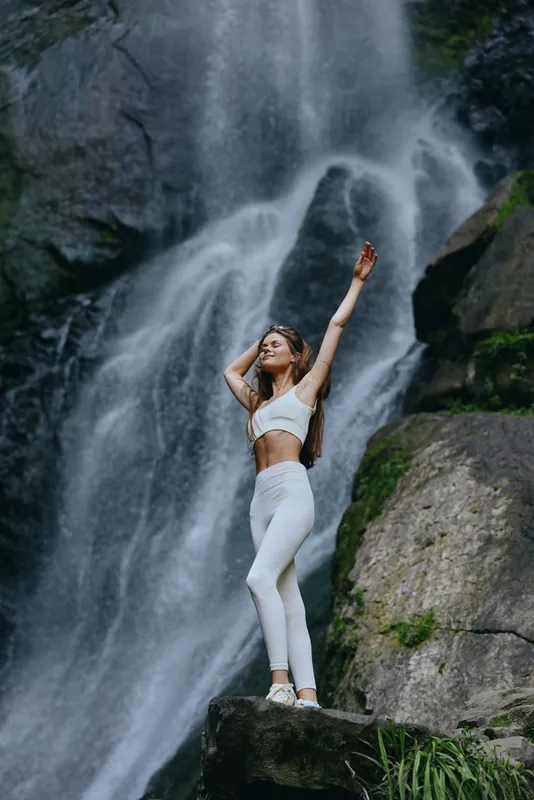 Young woman practicing yoga in white activewear stands confidently on a rock near a cascading waterfall surrounded by lush greenery and natural beauty.