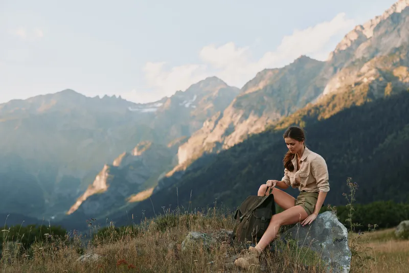 Young woman in casual clothes sits on rock with backpack in mountain valley enjoys nature and peaceful lifestyle during hiking trip under clear sky.