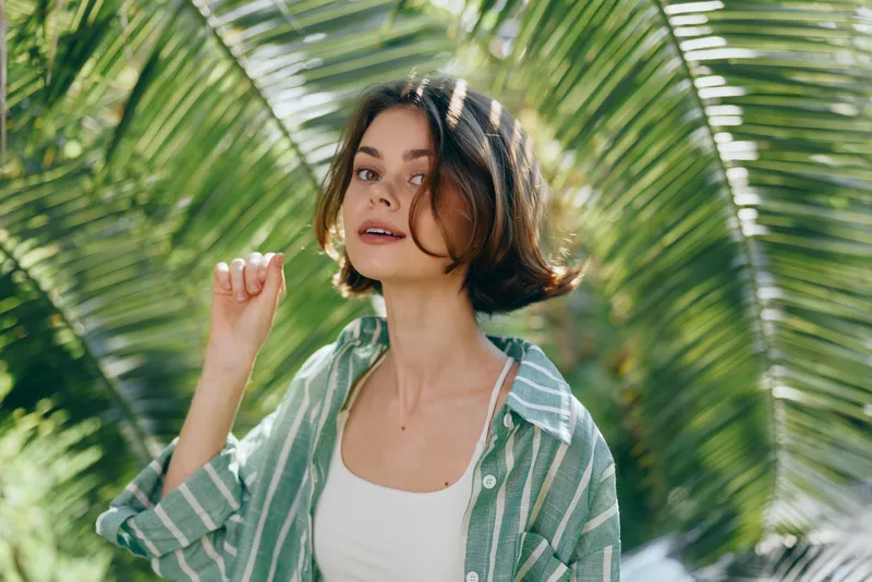 Portrait of a young woman outdoors among palm leaves, wearing a green striped shirt and white top, soft natural light.