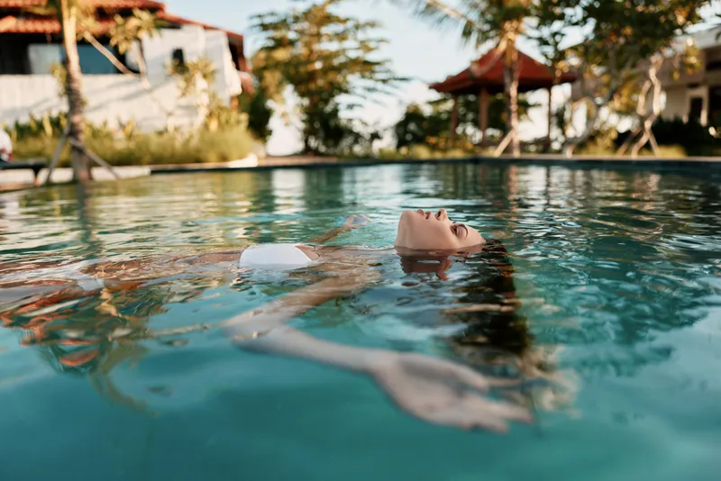 Person floating calmly in a clear pool under bright sun, capturing a relaxed outdoor scene by a sunny backyard. Wellness, leisure lifestyle moment with tranquil reflections and warm breeze.