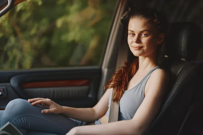 Young woman enjoying a relaxed drive in a stylish car, wearing a casual outfit and smiling warmly against a blurred green background