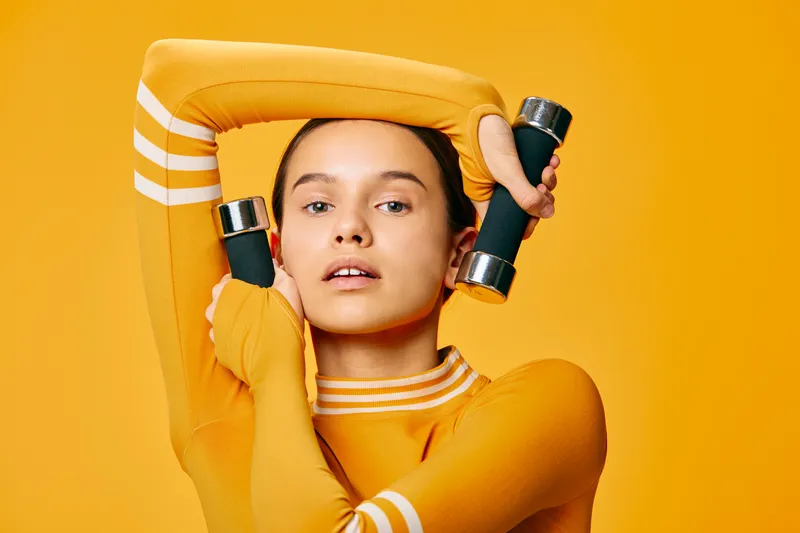 Athletic woman in a yellow sports outfit lifts small dumbbells in a studio with a bright orange backdrop, showcasing fitness and focus during a high-energy workout.