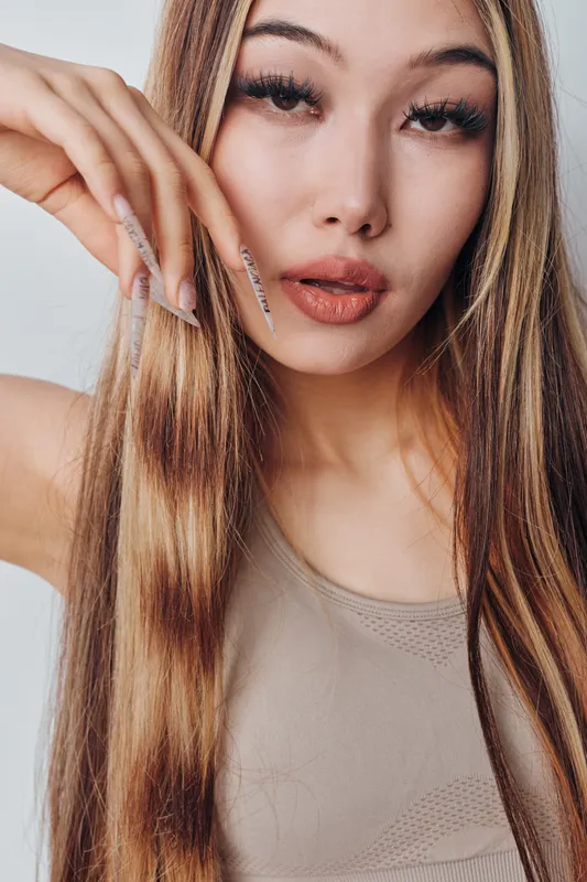 Young woman with long straight hair and manicured nails posing indoors. Closeup portrait with natural makeup and smooth skin wearing beige top in soft lighting.