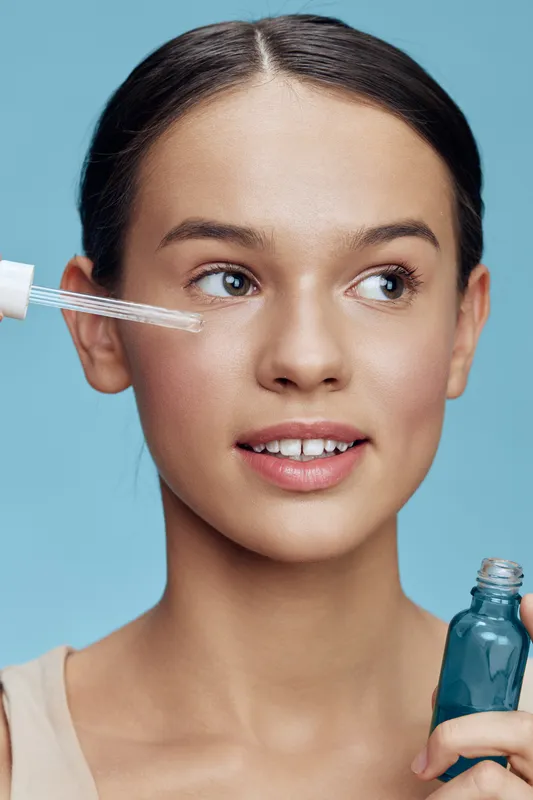 Young woman applying facial oil, promoting skincare routine with bright blue background, focused on healthy skin and beauty products Natural glow, clear complexion