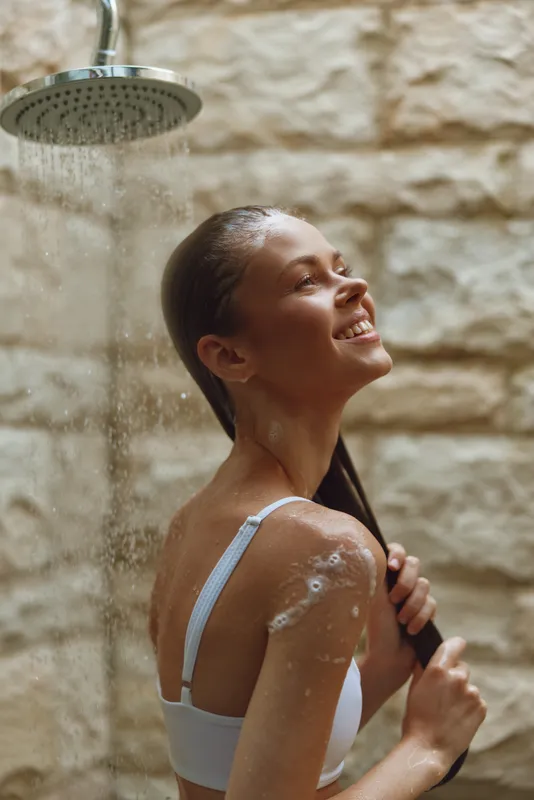 smiling woman enjoying warm outdoor shower with flowing water against textured stone wall, relaxed and refreshed mood in natural light