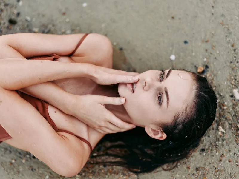 Closeup of a woman lying on a sandy beach, hair spread, skin illuminated by sun and water, calm gaze, and gentle pose conveying softness and vulnerability.