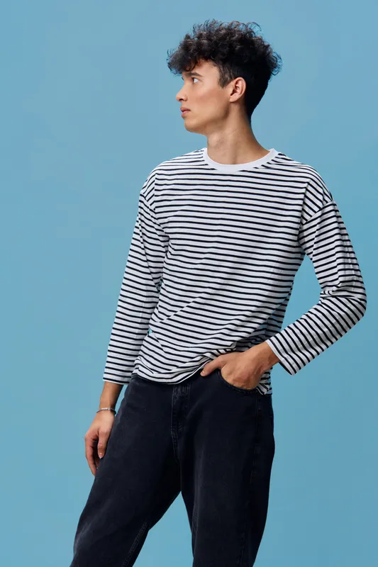Young man in striped long sleeve shirt and black pants stands against blue background looking sideways, casual style, curly hair, modern fashion portrait.