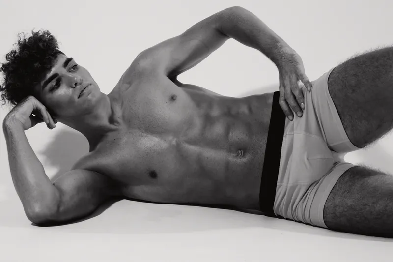 Young muscular man lying on side with hand behind head and wearing shorts in studio. Fitness model posing with toned body and relaxed expression on white background.
