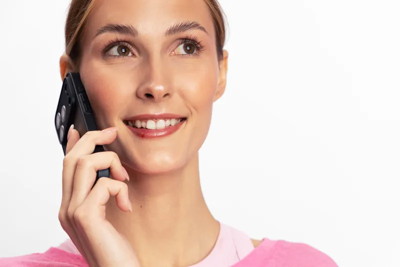 Smiling young woman with clear skin talking on mobile phone wearing pink top. Isolated on white background. Concept for communication, technology, and positive emotions.