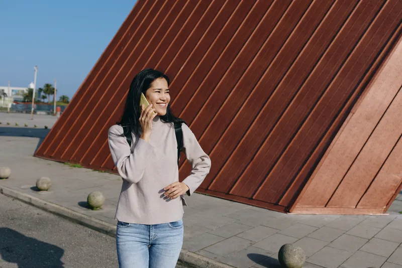 Woman talking on a phone outdoors near a bold diagonal red wall, casual outfit, smiling, urban street scene, everyday moment, travel vibe, openair architecture and sunlight.