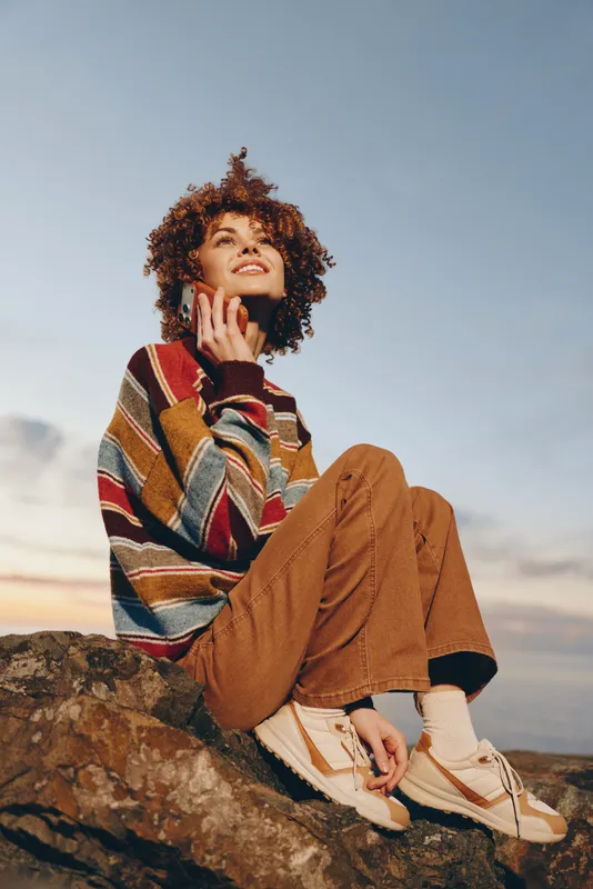 Woman lifestyle outdoor nature sunset scene, curly hair woman sitting on a rock, calm thoughtful expression, casual sweater and jeans, warm evening light, distant landscape