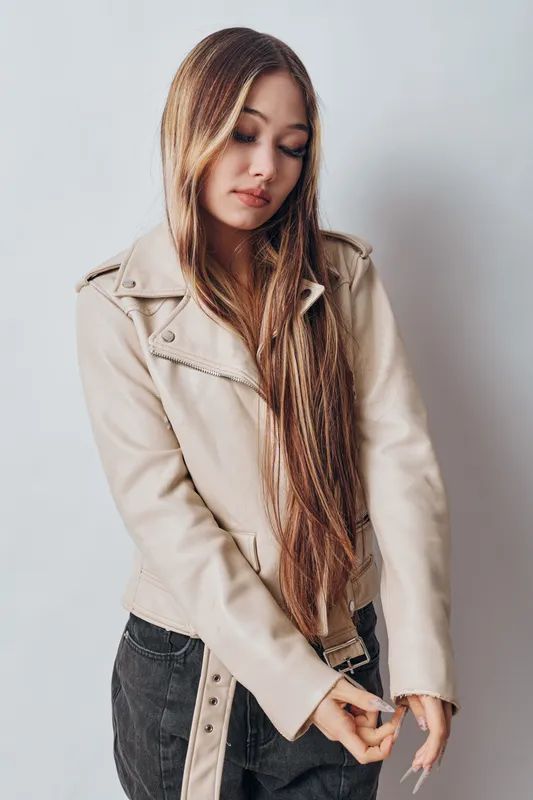 Young woman with long braided hair wearing beige leather jacket and black pants standing and looking down on white background. Stylish casual fashion portrait.