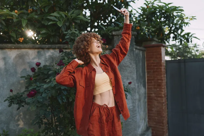 Woman with curly hair stands outdoors, wearing a red jacket and matching outfit, looking up and smiling as she raises her arm, framed by greenery and a brick wall, capturing a confident lifestyle